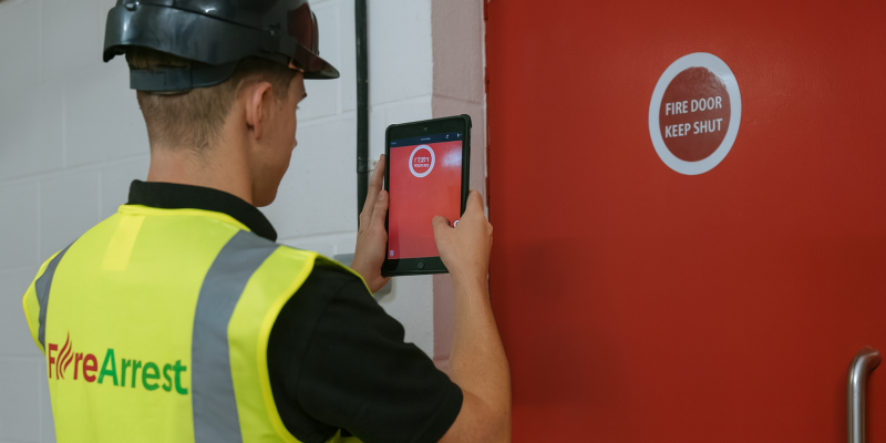 A fire safety inspector wearing a high-visibility vest and hard hat uses a tablet with a fire door inspection app to photograph a red fire door marked with a "FIRE DOOR KEEP SHUT" sign.
